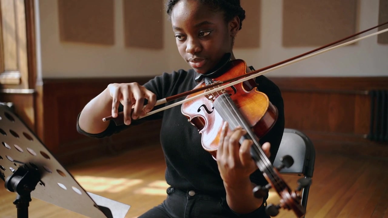 Glancing at score, violinist in black top, dark pants placing fingers, bowing violin at practice