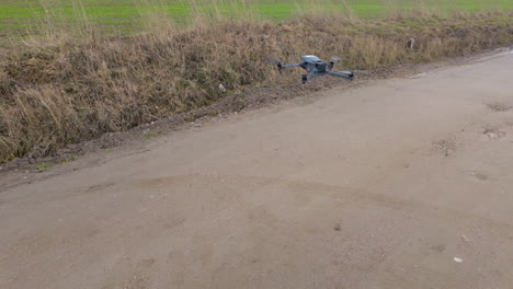 un dron con un cuerpo gris y luces azules está volando bajo sobre un camino de tierra, con hierba seca y una pendiente en el lado