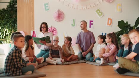 blonde girl in glasses with a bob hairstyle reads a green book for preschool. children children sit in a circle and listen carefully to their teacher. the second teacher, a man in a purple t-shirt, supports and listens attentively to the teacher