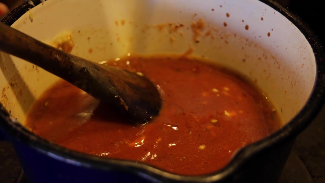 Primo piano a mano di salsa barbecue Cajun mescolata con un mestolo in una pentola blu sotto una fonte di luce al tungsteno