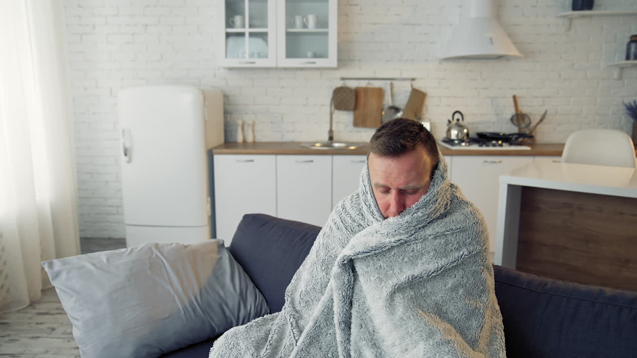 Man suffering from flu. Young sick man sitting on a sofa warming with blanket in the kitchen. Miserable man having a headache. Covid-19. Isolation at home.
