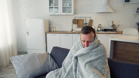 man suffering from flu. young sick man sitting on a sofa warming with blanket in the kitchen. miserable man having a headache. covid-19. isolation at home.