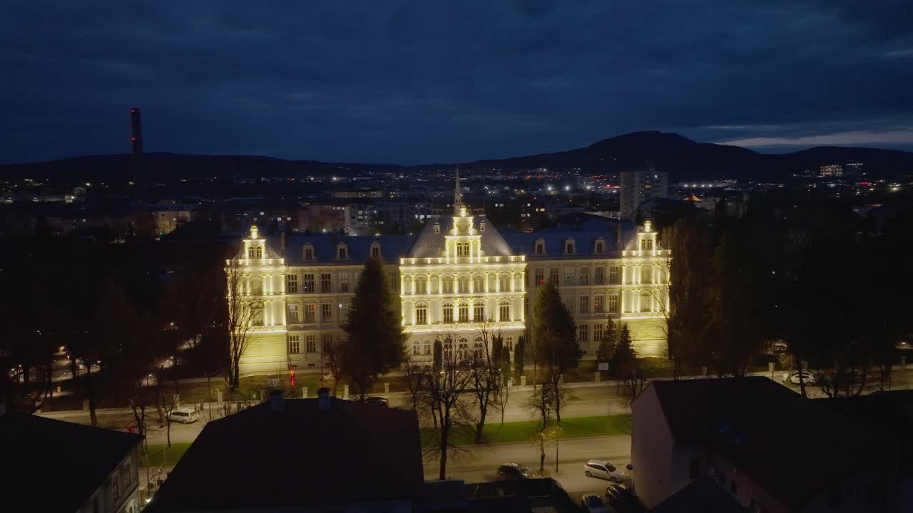 A wide aerial shot of the historic Liviu Rebreanu College in Bistrita, Romania. The grand building is beautifully illuminated against the twilight sky and city lights