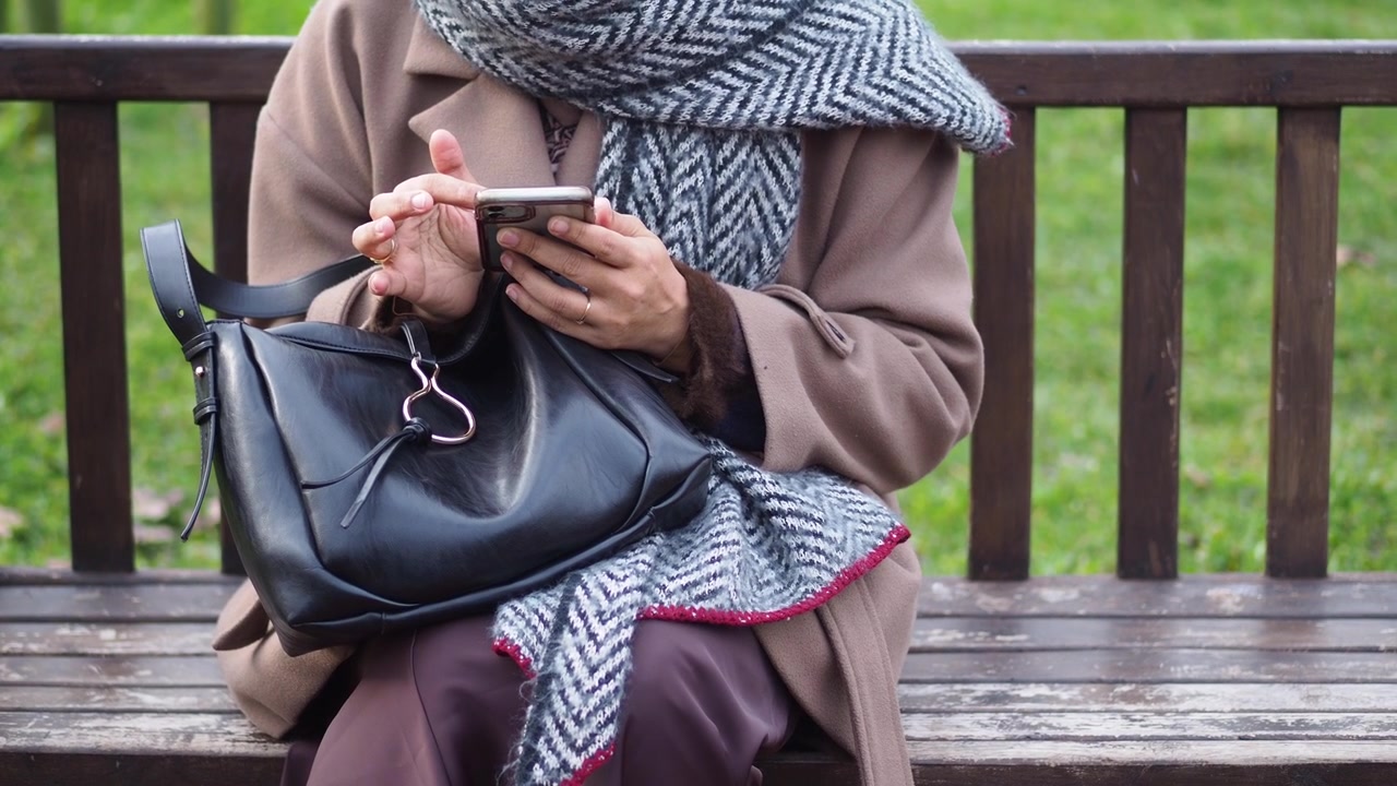 Kvinne i frakk og skjerf bruker smarttelefon på en parkbenk