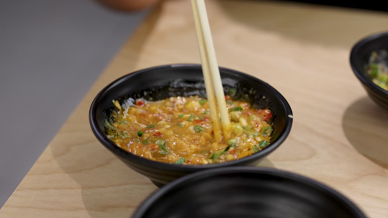 Hand stirs vibrant chili garlic sauce in black bowl using chopsticks, under soft, even lighting