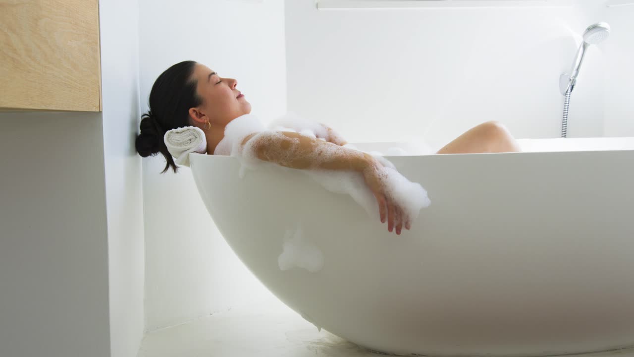 After settling into freestanding tub, woman is slowly sliding forearm and brushing foam for comfort