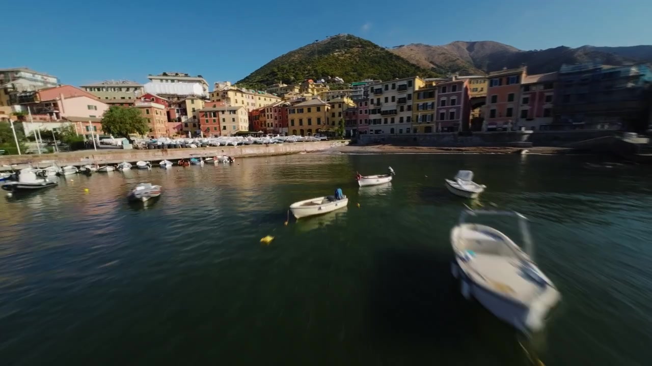 Volo dinamico di drone FPV su un piccolo porto con barche ed edifici colorati sul lungomare.
