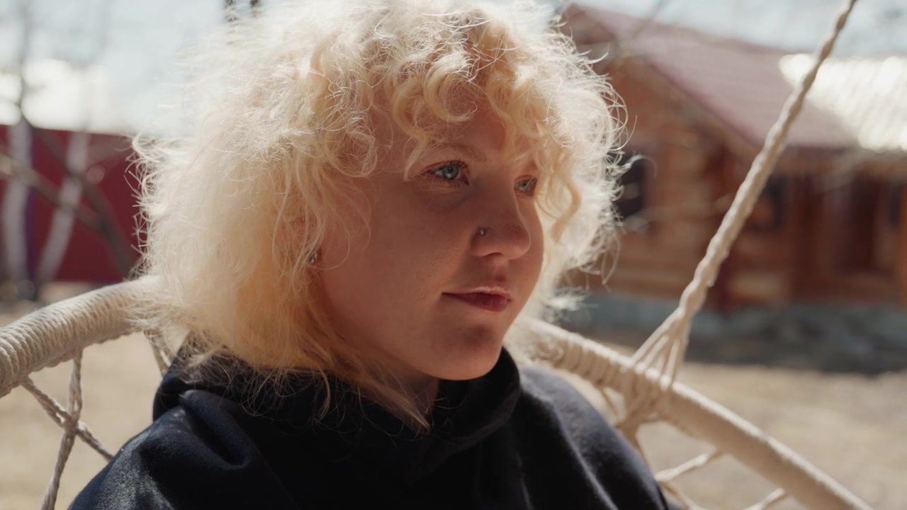 Nahaufnahme von Kopf und Schultern einer lockigen blonden Person, die in einem hängenden Korbsessel im Freien sitzt, sich leicht dreht mit Blick nach vorne, während der Wind die Locken anhebt; warme Mittagssonne hinterleuchtet Haare und weich.