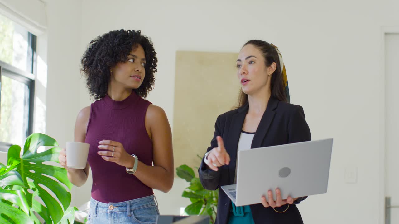 Collaboratrices diverses debout près de la fenêtre du bureau, consultant un ordinateur portable, tenant une tasse, vérifiant la lumière.