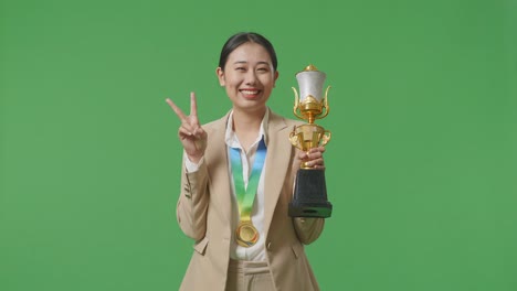 asian business woman in a suit with a gold medal and trophy showing peace gesture and smiling to camera as the first winner on green screen background in the studio