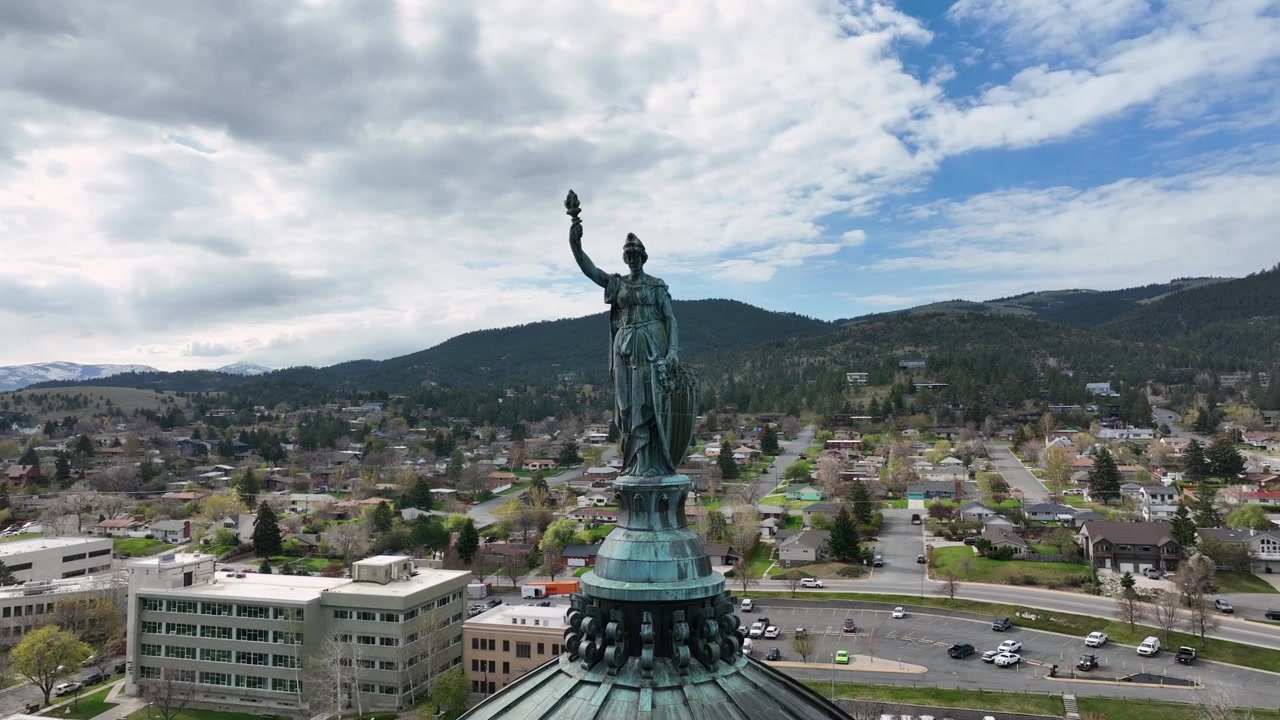 Statua di bronzo con torcia in cima a un pinnacolo di cupola con verderame, quartiere residenziale che si estende sotto verso montagne boscose e un moderno edificio per uffici. Helena, Montana, USA. Ripresa aerea con drone in orbita.