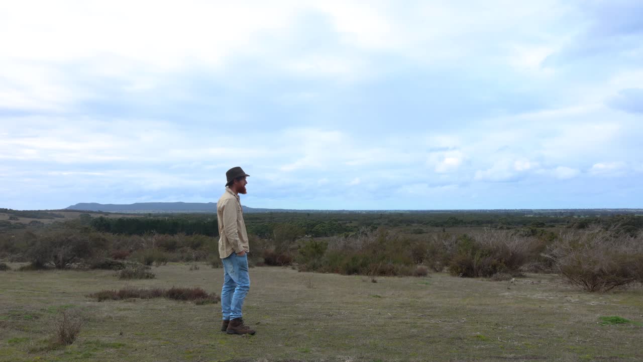 A bushman in a hat standing and looking out accross the Australian mallee desert in the green winter