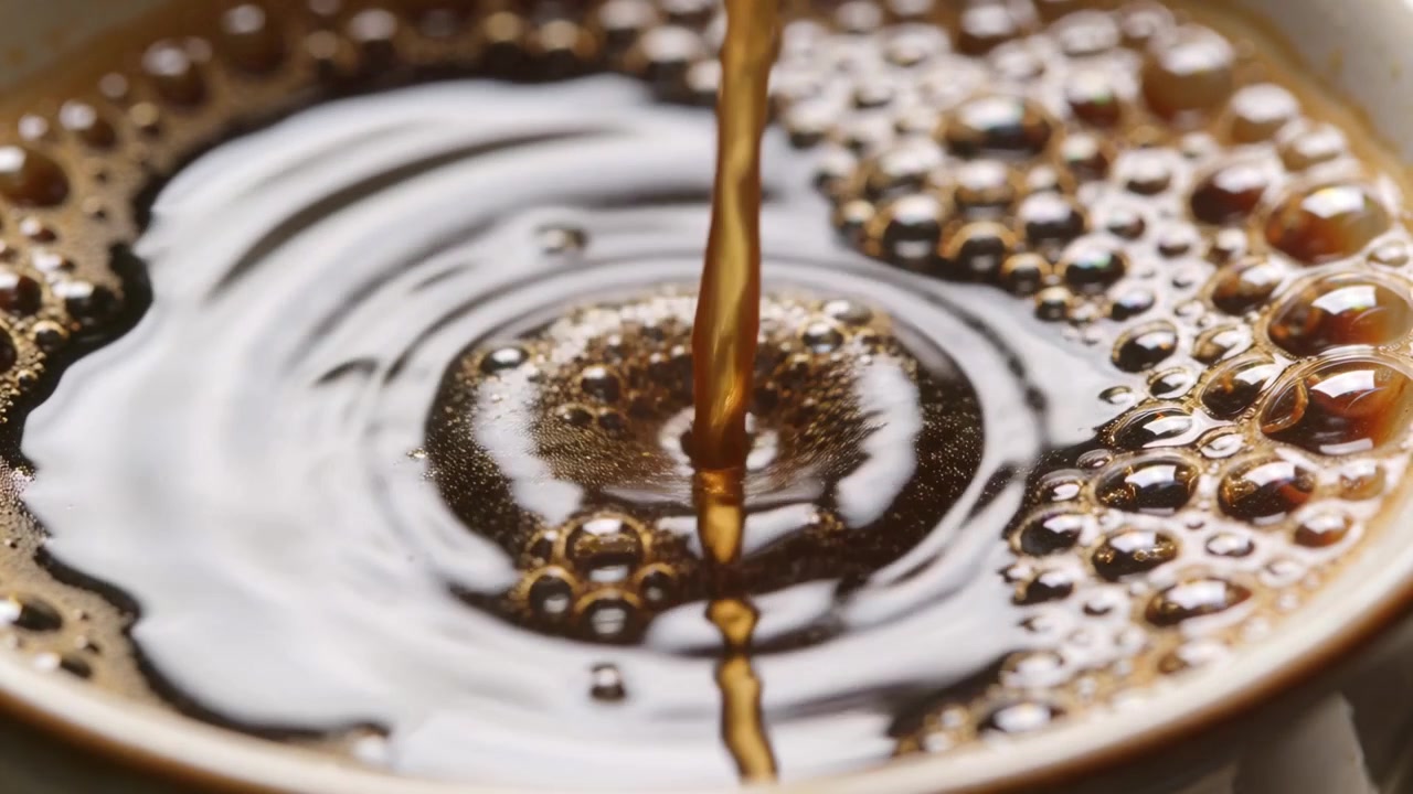 Kedel hælder mørk kaffestrøm ned i keramisk kop på køkkenbordet, fyldes med bobler.