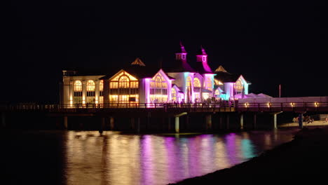 Timelapse-Nocturno-Del-Ascensor-Sellin-Pier-Iluminación-Colorida-Se-Refleja-En-Aguas-Tranquilas-Del-Mar,-Silueta-De-Turistas-Alemanes-Visitan-El-Balneario-En-La-Isla-De-Ruegen-Junto-Al-Mar-Báltico,-Alemania