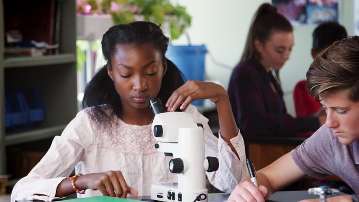 High School Students Looking Through Microscope And Writing Notes In Biology Class