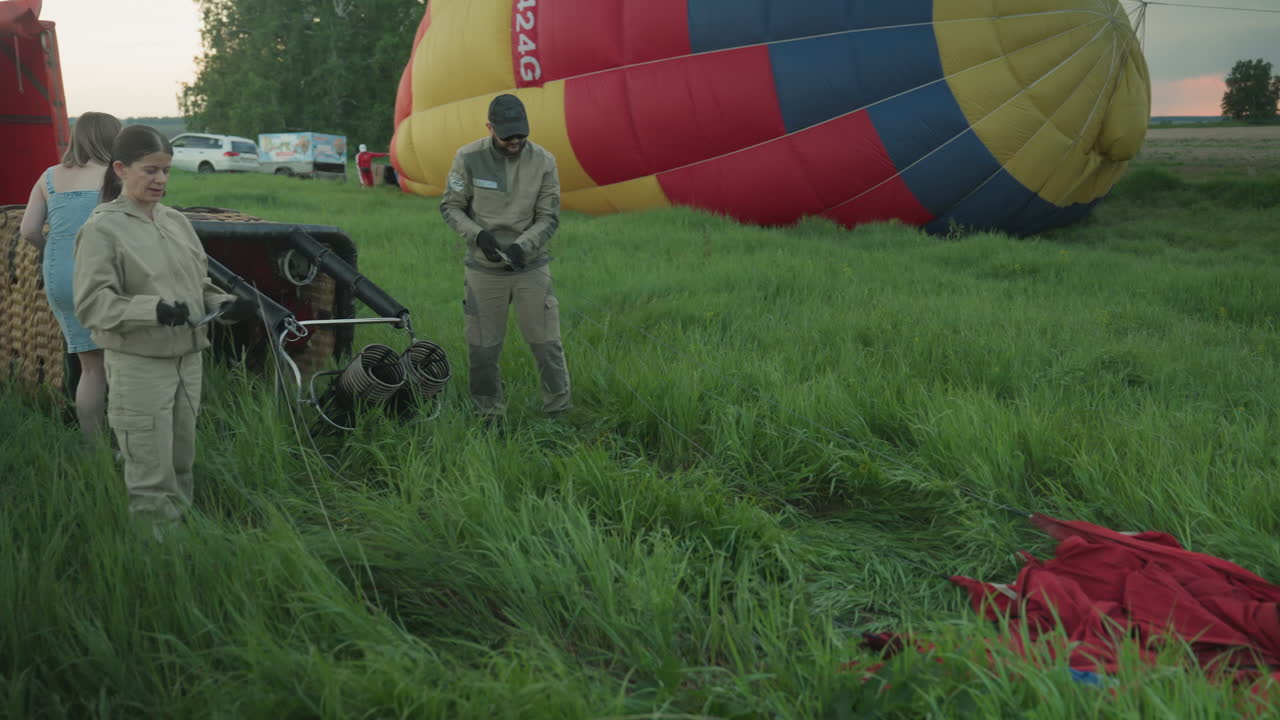 couple in matching jackets drag heavy red and green hot air balloon fabric across grassy field at dusk preparing envelope layout while sunset sky and distant trees