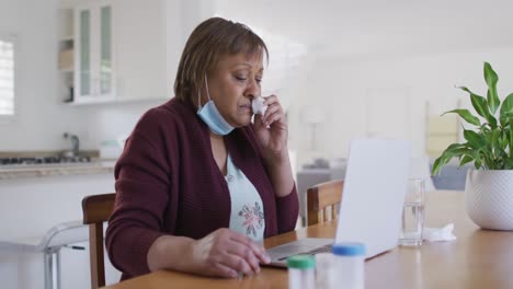 Sick-african-american-senior-woman-with-face-mask-using-laptop-at-home,-with-medication-on-table