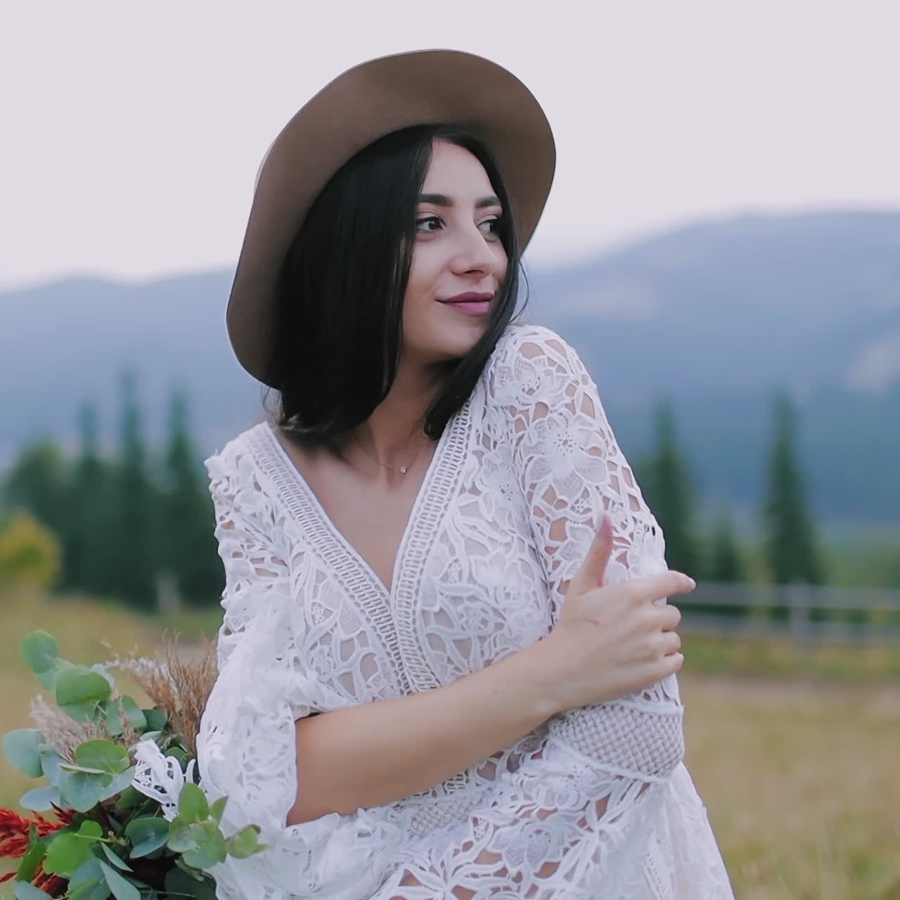Smiling bride in nature. Beautiful young woman in white dress and hat posing at camera on mountains background.