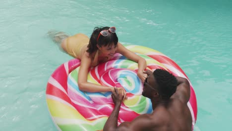 Happy-diverse-couple-wearing-swimming-suits-with-inflatable-at-swimming-pool-in-garden