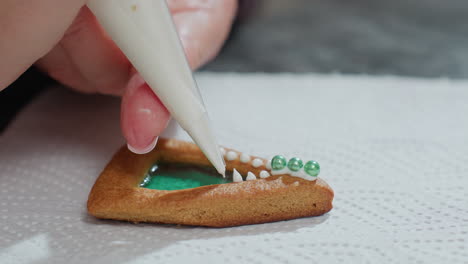 close-up of baker using white piping bag to decorate cookie with green color and white icing designs, baker focuses on precise detailing with beads and icing on the decorated surface