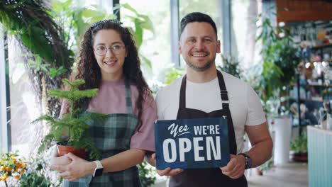portrait of girl and guy holding green plant and open sign in flower store