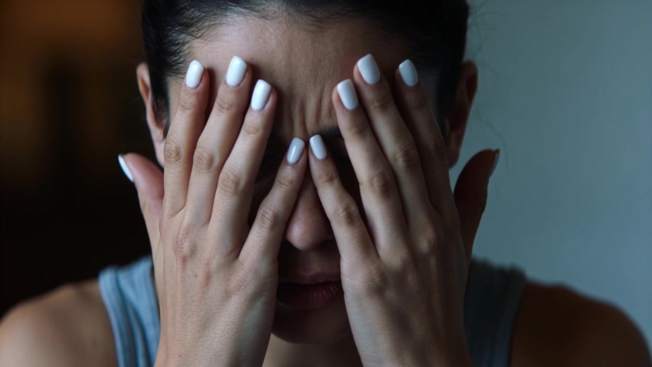 Donna accigliata si preme le tempie e poi si copre gli occhi in una stanza, sentendosi stressata, con unghie bianche e canotta grigia.