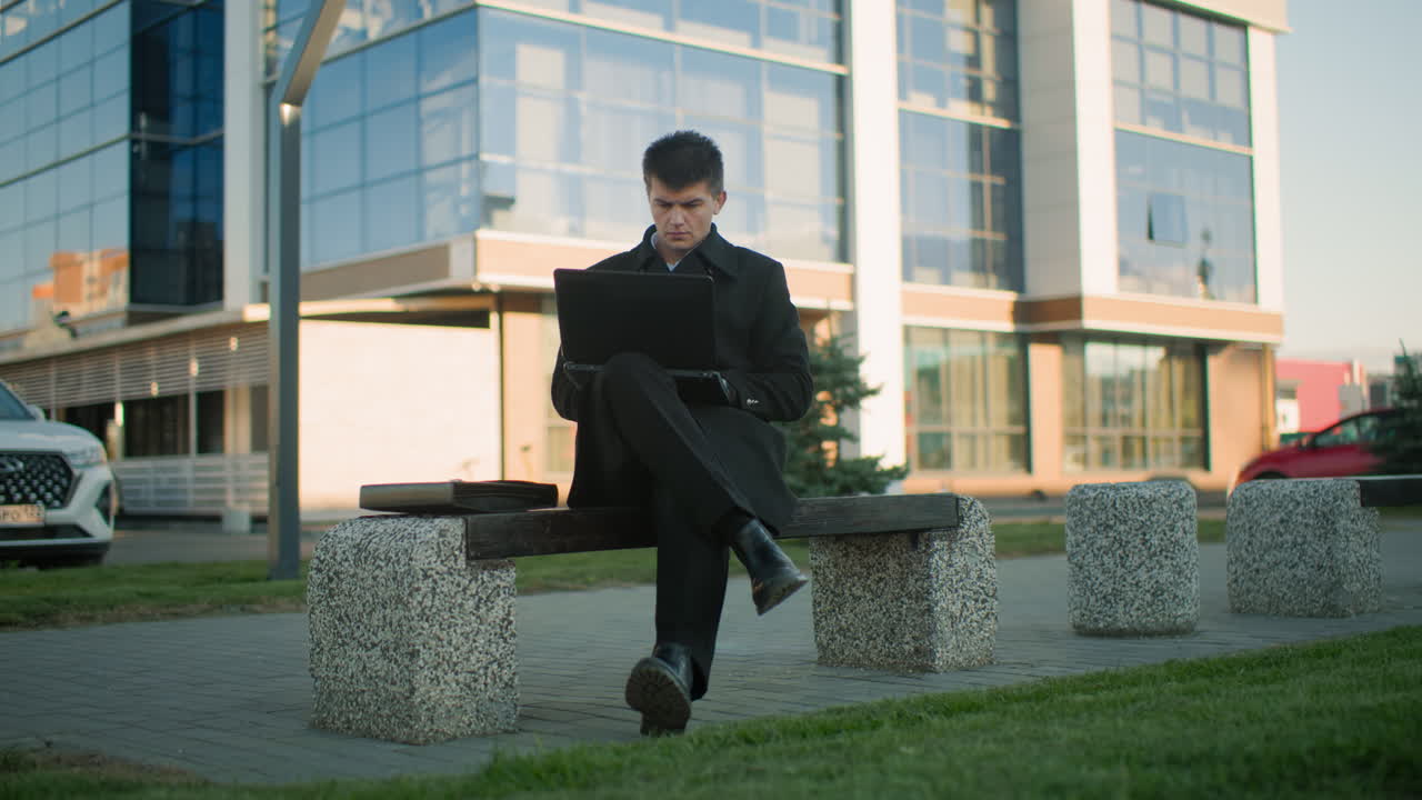 hombre de negocios sentado al aire libre trabajando en una computadora portátil con las piernas cruzadas en un entorno urbano moderno, el fondo incluye coches estacionados, un edificio contemporáneo con fachada de vidrio