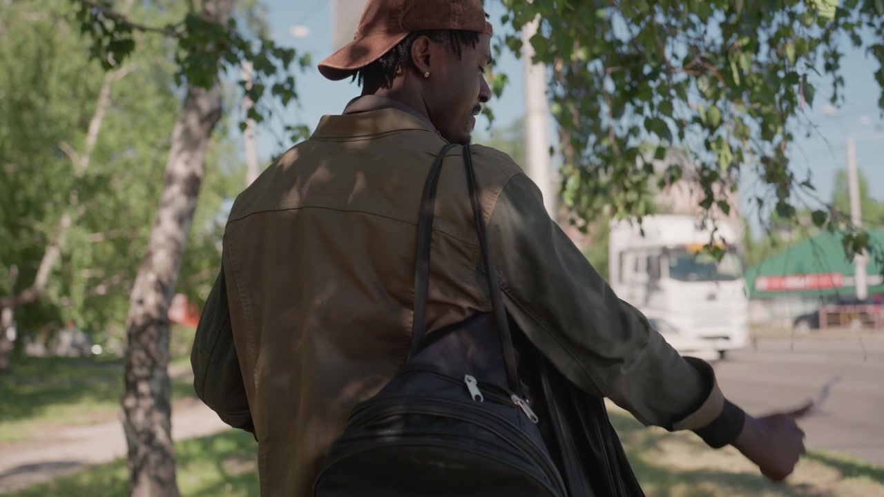 Man with backpack examines paper and gives thumbs up on roadside. Man wears olive jacket brown cap carries black bag holds map or document under tree lined street as vehicles pass