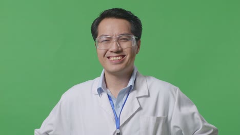 close up of asian man scientist with arms akimbo looking at camera and smiling while standing on the green screen background in the laboratory