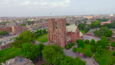 Vista-Aérea-De-Una-Hermosa-Iglesia-Antigua-En-La-Ciudad,-El-Tráfico-Se-Mueve-En-El-Otro-Lado-De-La-Calle-De-La-Iglesia,-Hermosa-Vegetación-Alrededor-De-La-Iglesia,-Una-Bandera-En-La-Parte-Superior-De-La-Iglesia