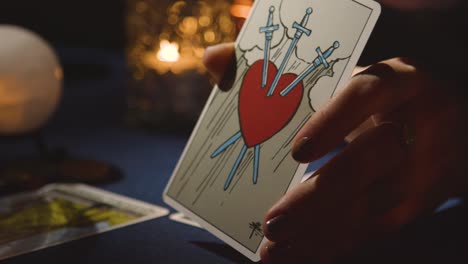 primer plano de una mujer dando lectura de cartas del tarot en una mesa a la luz de las velas sosteniendo una tarjeta que muestra un corazón atravesado por espadas 1