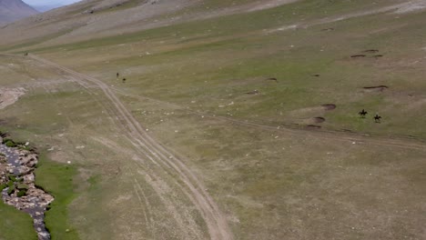 Traditional-nomadic-horsemen-of-mongolia,-horseriding-in-a-green-valley-landscape,-idyllic-mongolian-grassland-with-a-small-river-stream,-aerial-drone-overview-shot