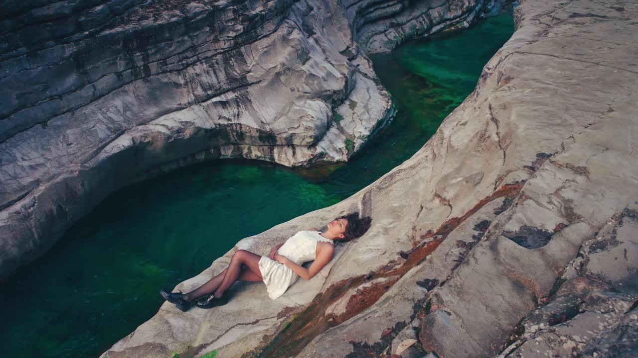4K Cinemagraph - seamless video loop of a young brunette model woman in a white dress lying by the edge of a gorge by a blue mountain river. The water is rushing through naturally formed rocks