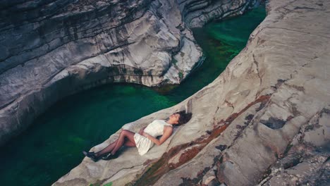 4k cinemagraph - seamless video loop of a young brunette model woman in a white dress lying by the edge of a gorge by a blue mountain river. the water is rushing through naturally formed rocks