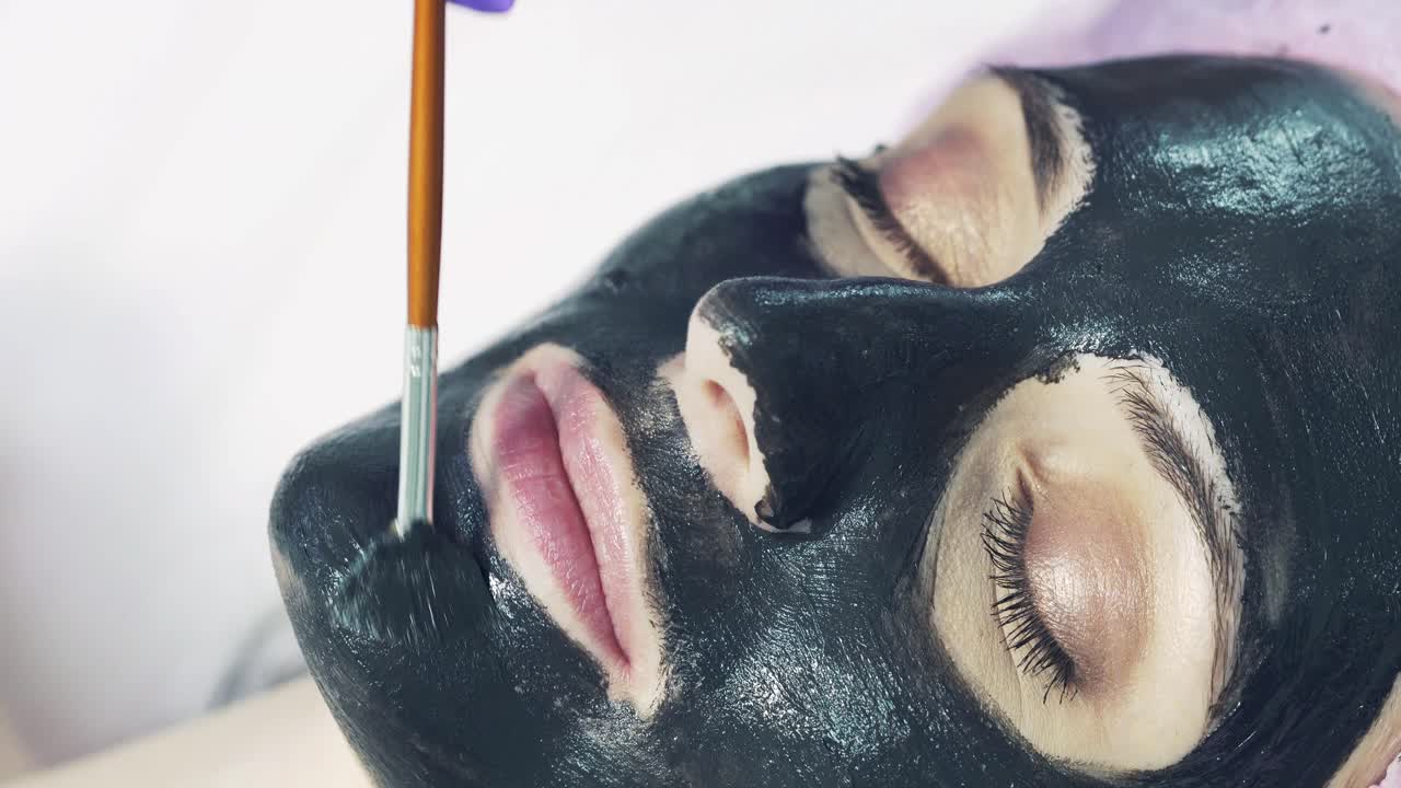 Beautiful woman is applying facial black mask at beauty salon. Spa treatment on face for young woman