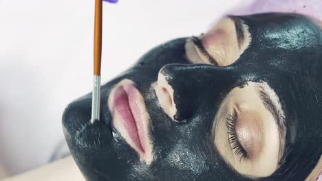 beautiful woman is applying facial black mask at beauty salon. spa treatment on face for young woman