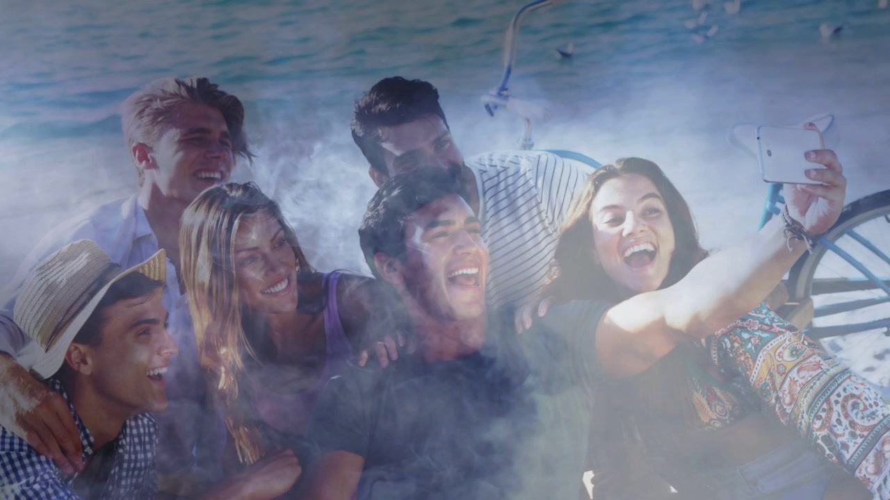 Pessoa à direita levantando o telefone, grupo se inclinando, rindo na praia, névoa cruzando rostos para marketing