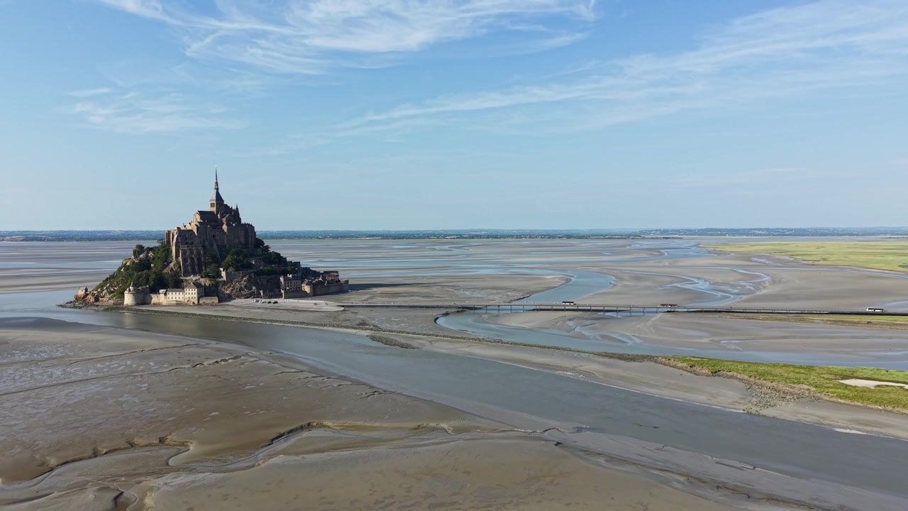 Plan aérien cinématographique par drone en recul du Mont Saint-Michel, révélant progressivement l'emblématique abbaye médiévale, de vastes vasières, des chenaux de marée sinueux et la chaussée reliant l'île au continent.