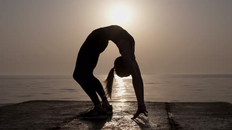 Female-silhouette-with-a-beautiful-morning-sky-and-calm-sea-ocean-on-the-background.-Slim-girl-exercising,-doing-bridge-pose.-Flexibility.-Pier.-Front-view