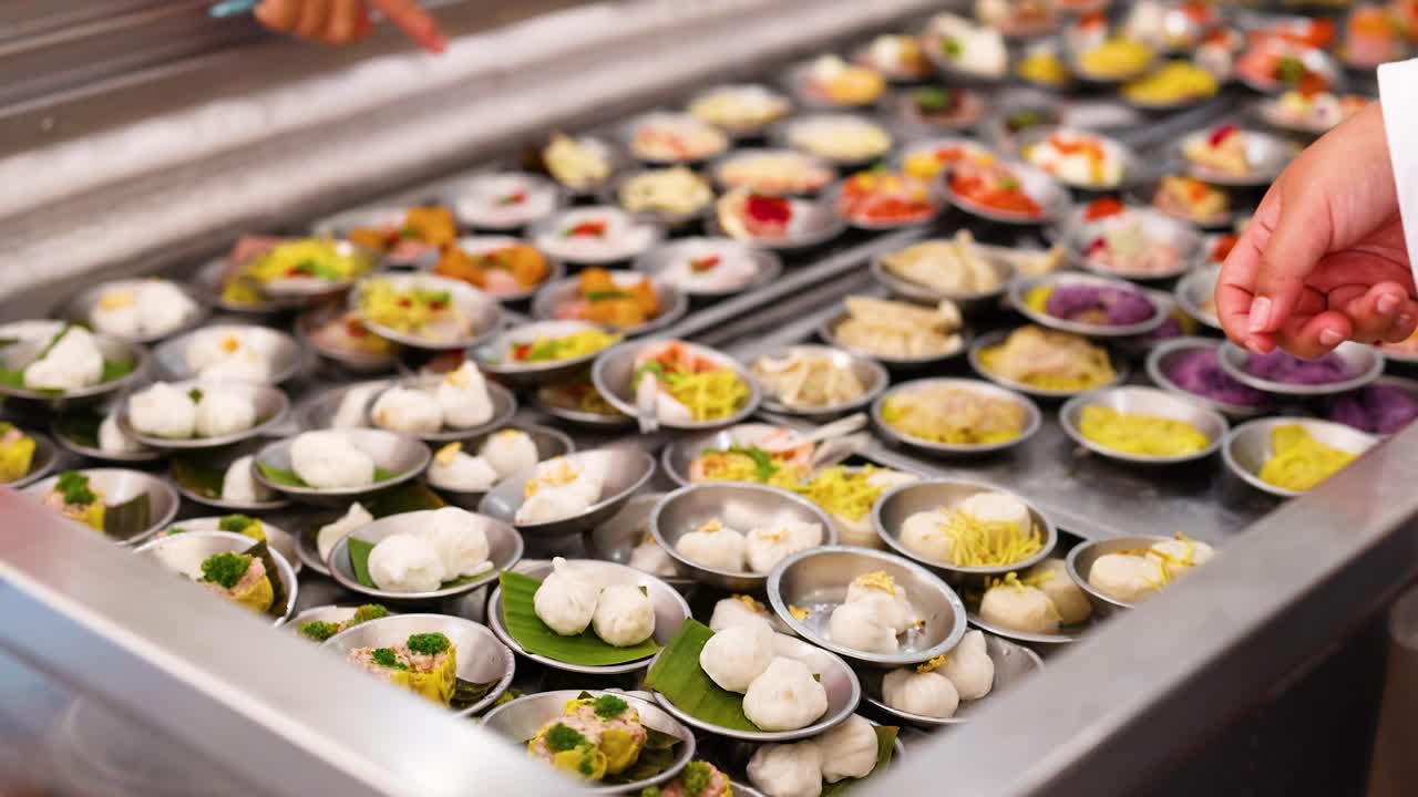 Hands choose dim sum from a vibrant array in a bustling Phuket market. Bright lighting highlights the colorful variety