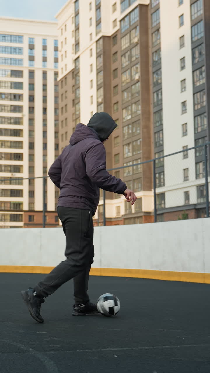 Young athlete skillfully controlling soccer ball with precise footwork during training session on sport arena, background includes urban buildings, fencing, and goalpost under soft daylight