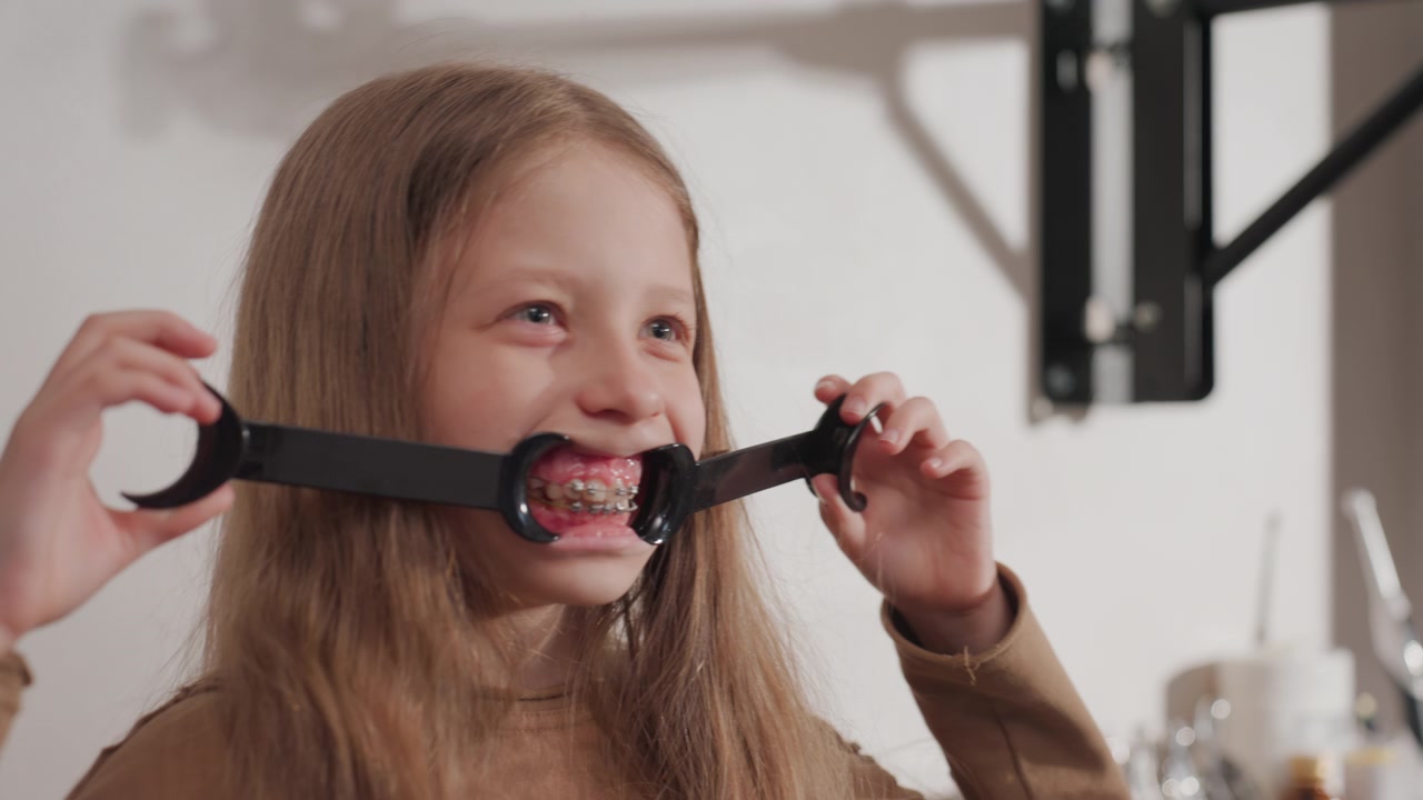 Primo piano di una giovane ragazza che tiene un divaricatore dentale nero per mostrare l'apparecchio ortodontico in bocca. La ragazza con i capelli lunghi indossa un top marrone, lo sfondo include attrezzature mediche e una parete bianca. Morbido.