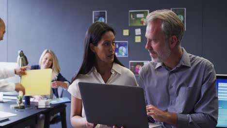 Diverse-business-people-using-laptop-talking-in-office-and-colleagues-working-behind