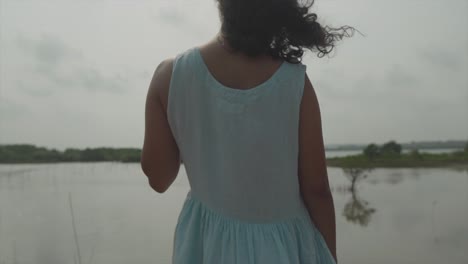 Toma-Panorámica-En-Cámara-Lenta-Media-De-Una-Mujer-India-Elegante-Vestida-De-Turquesa-Frente-A-Un-Lago-Con-Islas-Mientras-Su-Cabello-Sopla-En-El-Viento-Y-Mira-Hacia-El-Lago