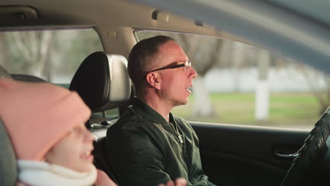 a young girl wearing a pink beanie and jacket is seated in the front passenger seat of a car, while a man, wearing glasses and a green jacket, is in the driver's seat, singing enthusiastically