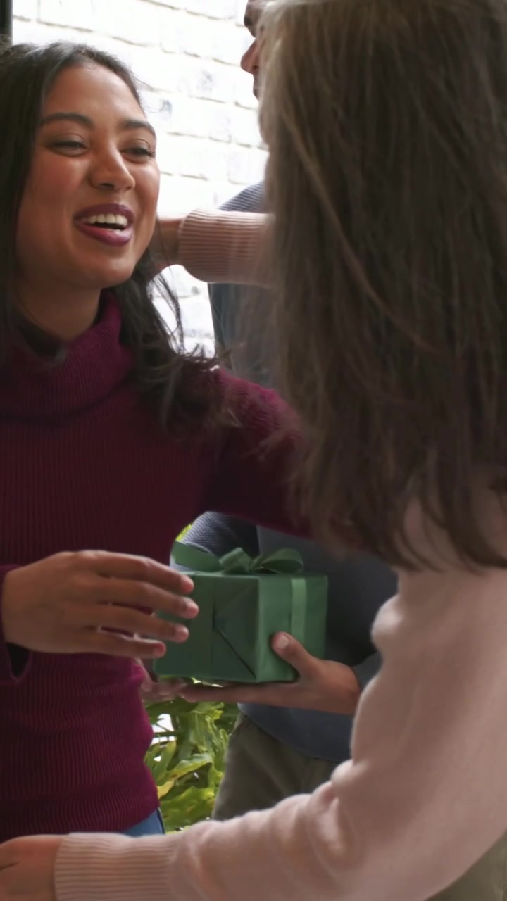 Video verticale: Un uomo arriva con un regalo verde, e una donna con un maglione rosa abbraccia un ospite alla porta.