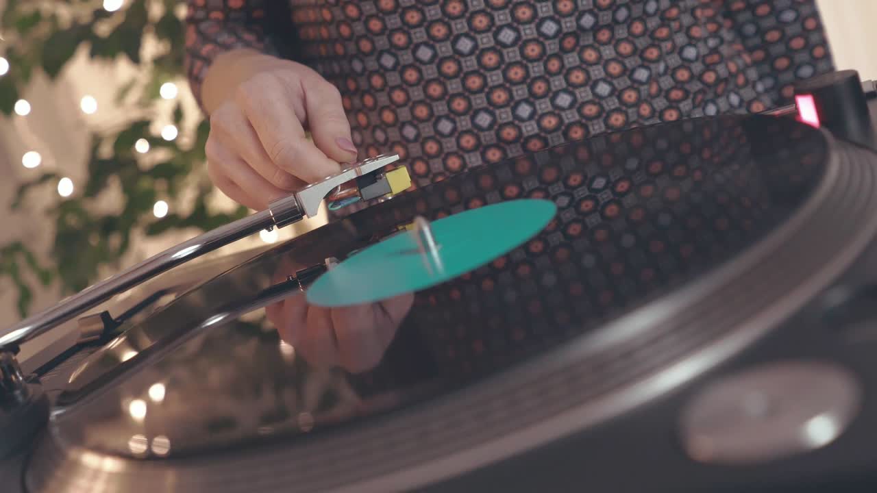 드레스를 입은 한 소녀가 dj 턴테이블 레코드 플레이어에 가까이 다가와 회전하는 검은 비닐 레코드에 바늘을 가진 스타일러스를 넣고 밝은 파티 조명을 배경으로 춤을 추고 으로 움직입니다.