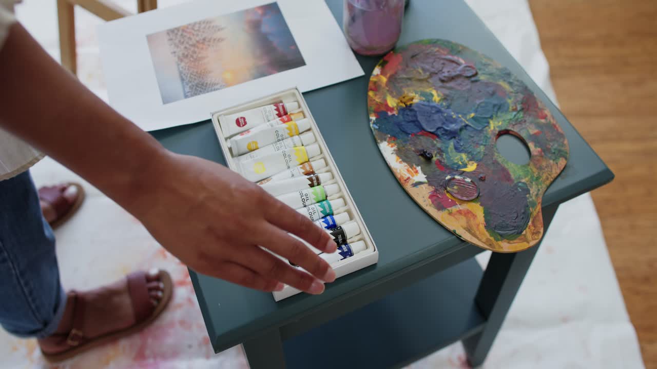 Donna afroamericana che si protende per ispezionare tubetti di pittura su un tavolo color ottanio che si abbina a un vasetto per tavolozza di stampa