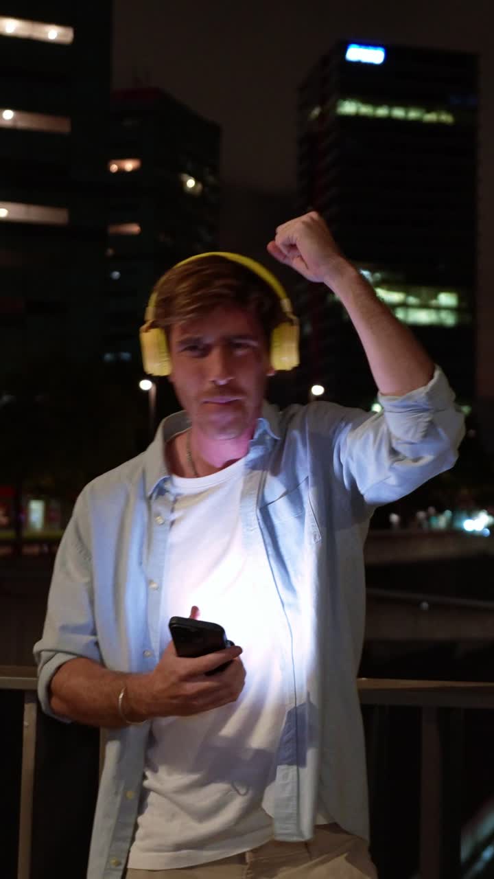 Giovane uomo entusiasta con le cuffie festeggia il successo sullo smartphone di notte