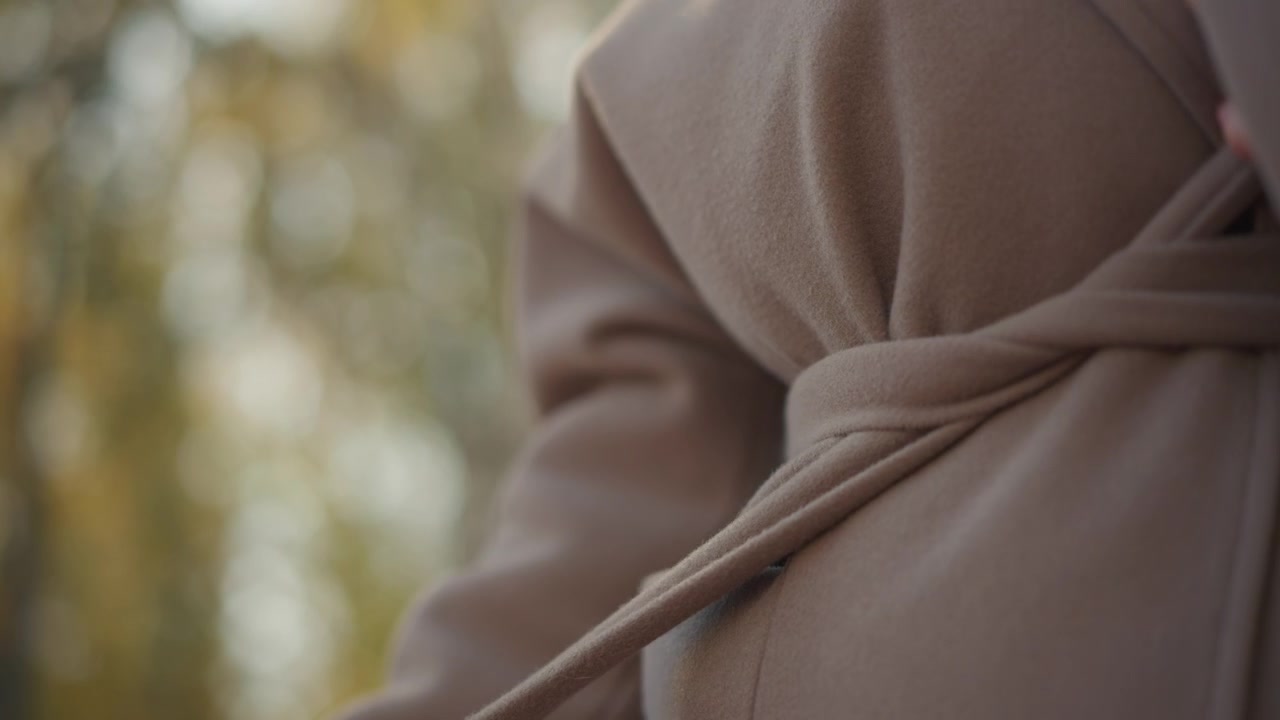 Close up of woman adjusting belt on her brown coat focusing on hands and fabric texture. subject hand moves to grasp or smooth material as woman interacts with clothing details. Soft